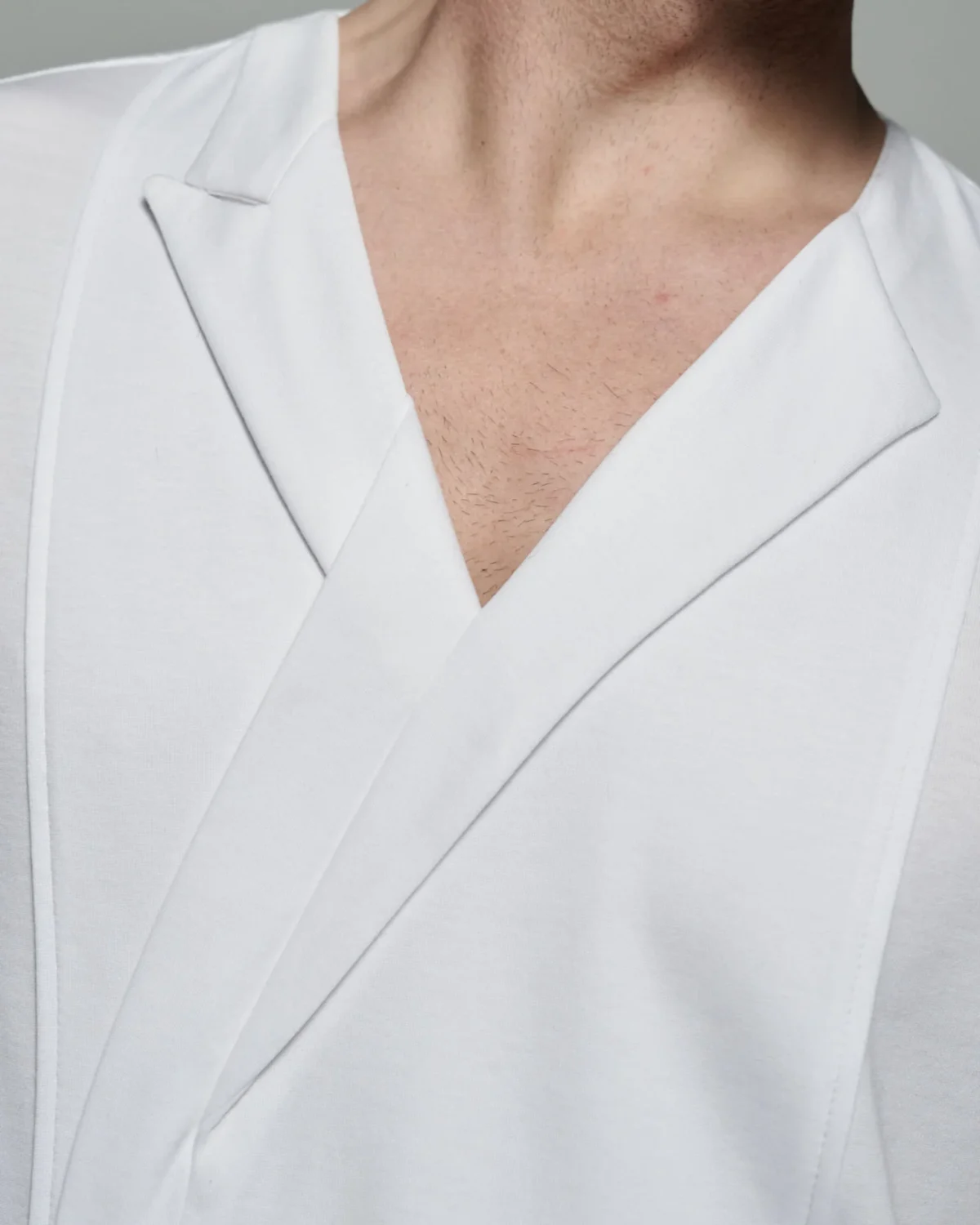 This image features a close-up of the W. Rubens 73, an Asymmetric V-neck T-shirt with Lapels & Inserts, elegantly showcasing its unique design elements. The T-shirt, crafted in England, highlights the brand's commitment to redefining conventional style through innovative design. The V-neckline is framed by asymmetric lapels, where a peak lapel graces the left side, offering a structured and refined appearance, while the right side exhibits a collarless design, creating an avant-garde and contemporary look. The image also reveals the artful folded insert on the front body panel, a distinctive feature that enhances the T-shirt's aesthetic and provides a sense of dimension and texture. The contrast between the soft, smooth white cotton fabric and the precise grey top stitching adds a touch of finesse, elevating the overall design. This stitching not only enhances the visual appeal but also ensures the garment's durability and longevity. The slim block fit, as seen in the image, is designed to contour the body without being restrictive, offering a comfortable and flattering silhouette. The fit is further refined by the French side seams, which maintain the T-shirt's polished appearance and ensure that it holds its shape over time. The choice of fabric and craftsmanship ensures that this T-shirt feels luxurious against the skin, offering both comfort and style.