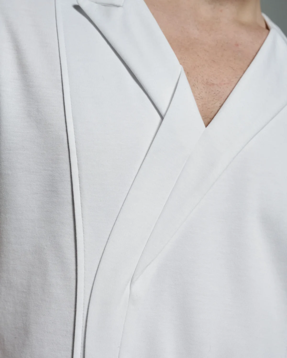 The W. Rubens 73 white T-shirt showcases an asymmetric V-neck with lapels and inserts, masterfully tailored in London. The close-up highlights the elegant structure of the lapels, with clean lines that emphasize the minimalistic design. The soft cotton fabric drapes naturally on the chest, demonstrating the shirt's luxurious craftsmanship. The distinctive collar and sharp angles represent W. Rubens' commitment to modern British artistry.