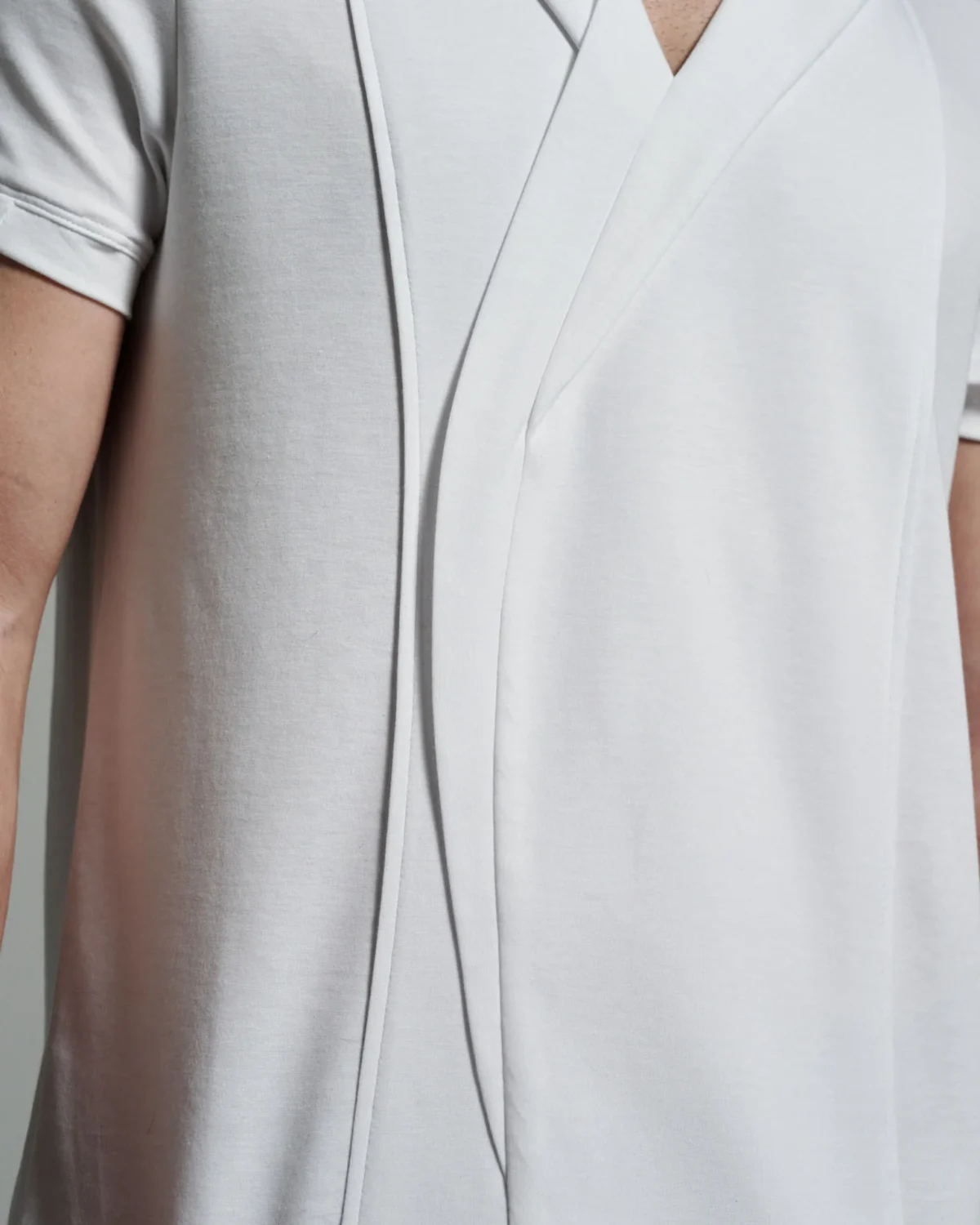 A full view of the W. Rubens 73 white T-shirt with asymmetric V-neck lapels and inserts, offering a glimpse of its contemporary design. The shirt's fitted silhouette, shown on a model, accentuates its sleek cut. The asymmetric lapels cascade down the front, creating a sophisticated visual effect that reflects the meticulous attention to detail in British-made garments. The fabric’s natural stretch ensures a flattering fit while maintaining comfort.